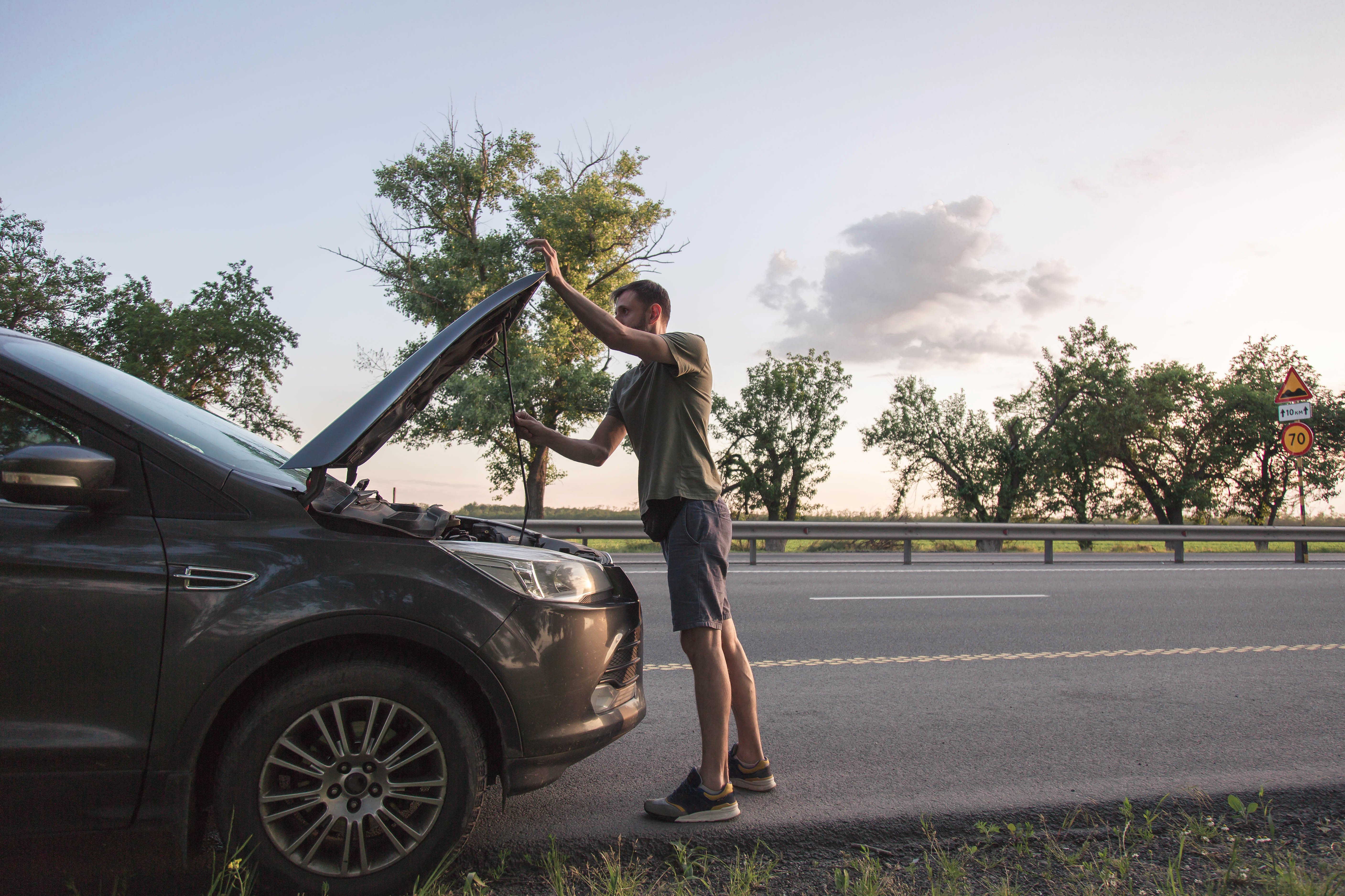 Man inspecting broken car with open hood on roadside highway at sunset, vehicle breakdown during road trip, transportation problem concept, emergency assistance, automotive repair, travel disruption